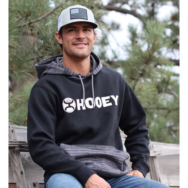 male model sporting the lock-up hoody in black with heather grey pocket and hood, grey and white liberty roper cap and jeans seated near a scrap wood pile